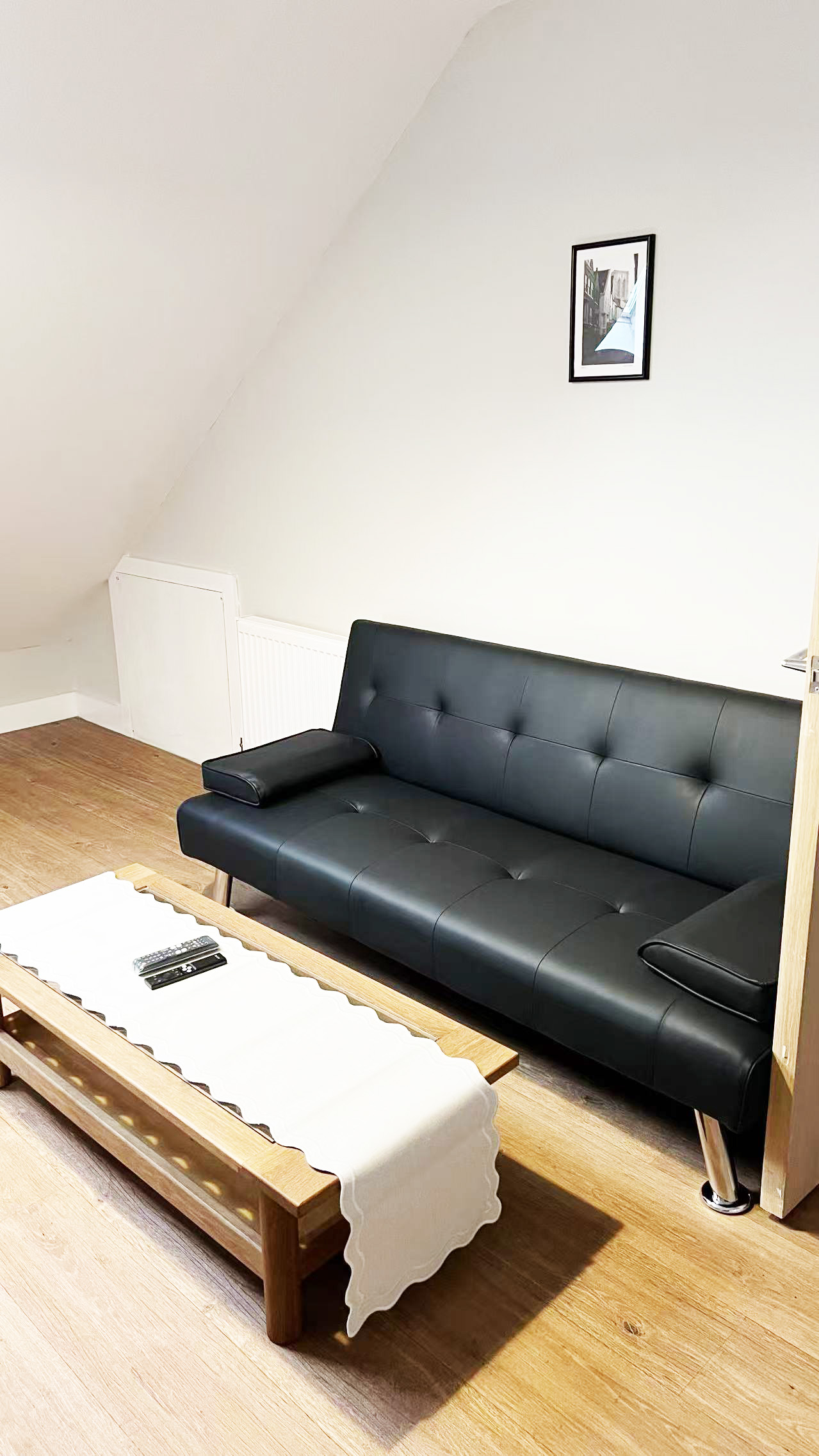 Small living room with a black tufted leather sofa against a slanted white wall, a wooden coffee table with a white runner, and a framed picture on the wall.