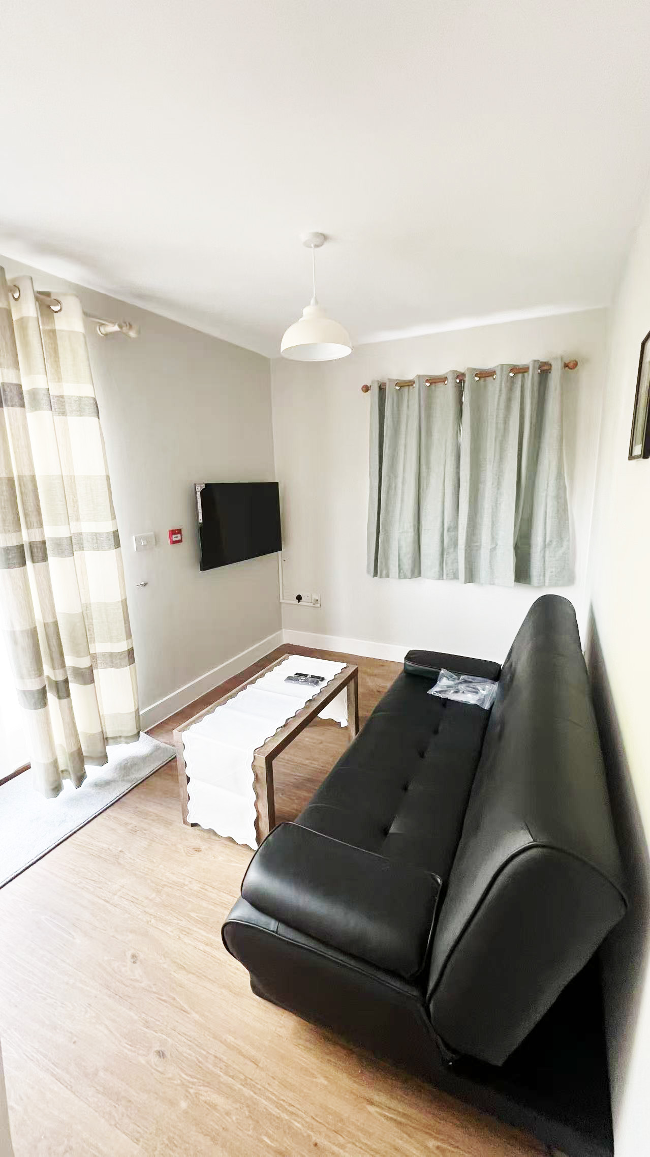 Living room with a black leather sofa, light wood floor, and a rectangular coffee table; wall-mounted TV on a gray wall beside striped curtains and a window.