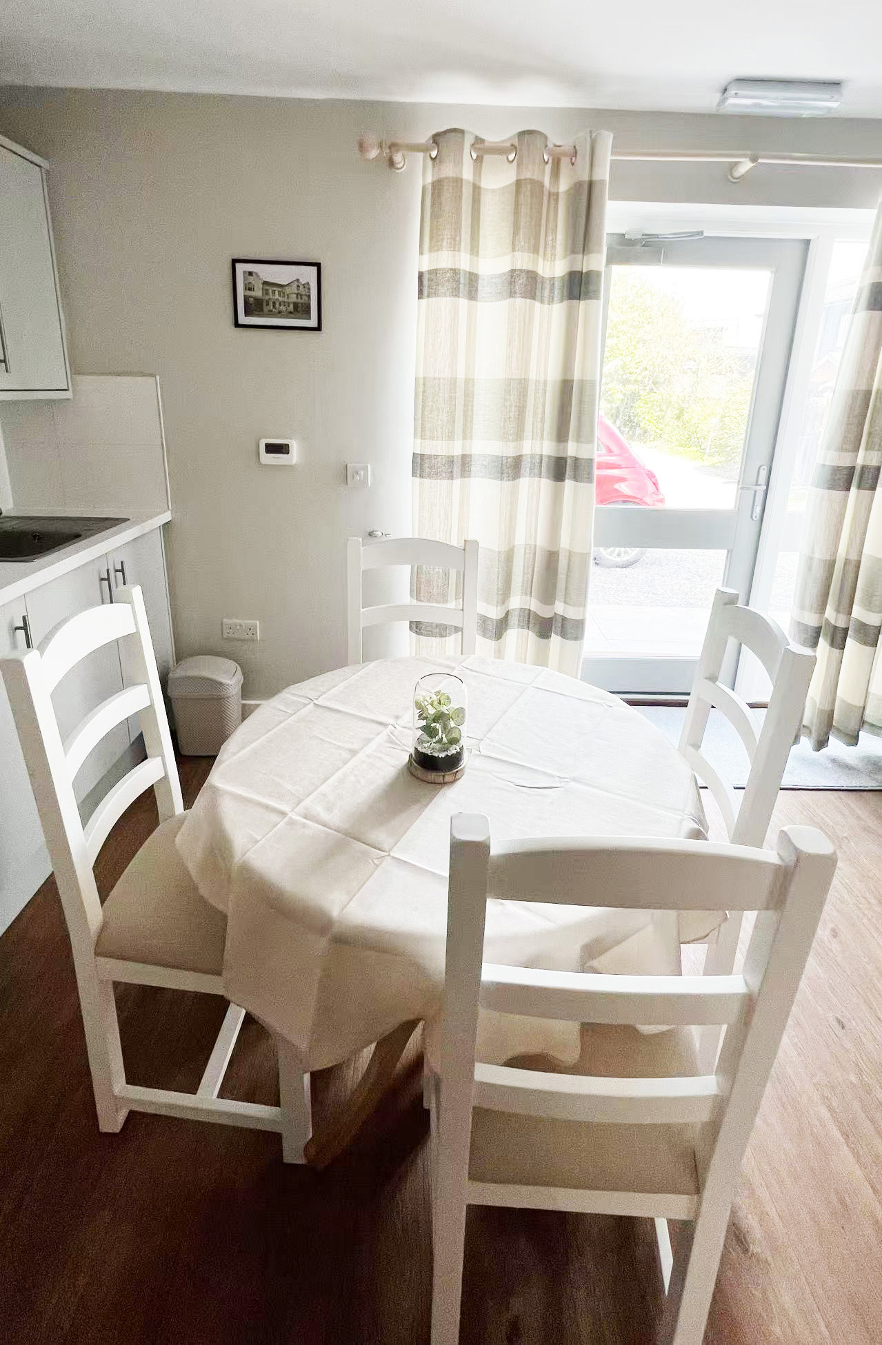 Round dining table with a white tablecloth and white chairs in a bright kitchen nook; a small glass terrarium centerpiece sits at the center, with striped beige curtains and a glass door to the outside where a red car is visible.