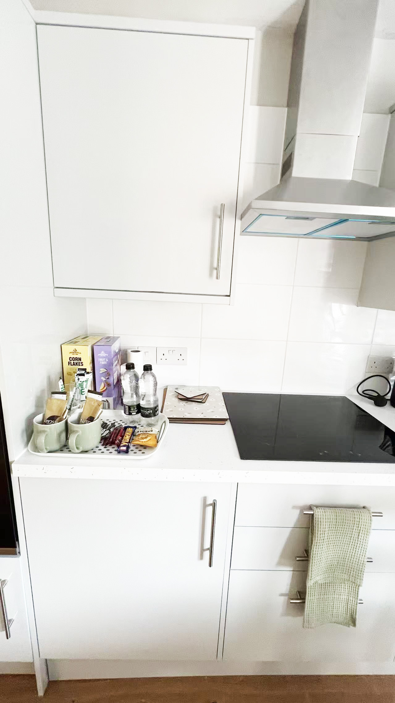 Modern white kitchen with an induction hob and stainless range hood; countertop holds mugs, snacks, and two water bottles near cereal boxes on the left.
