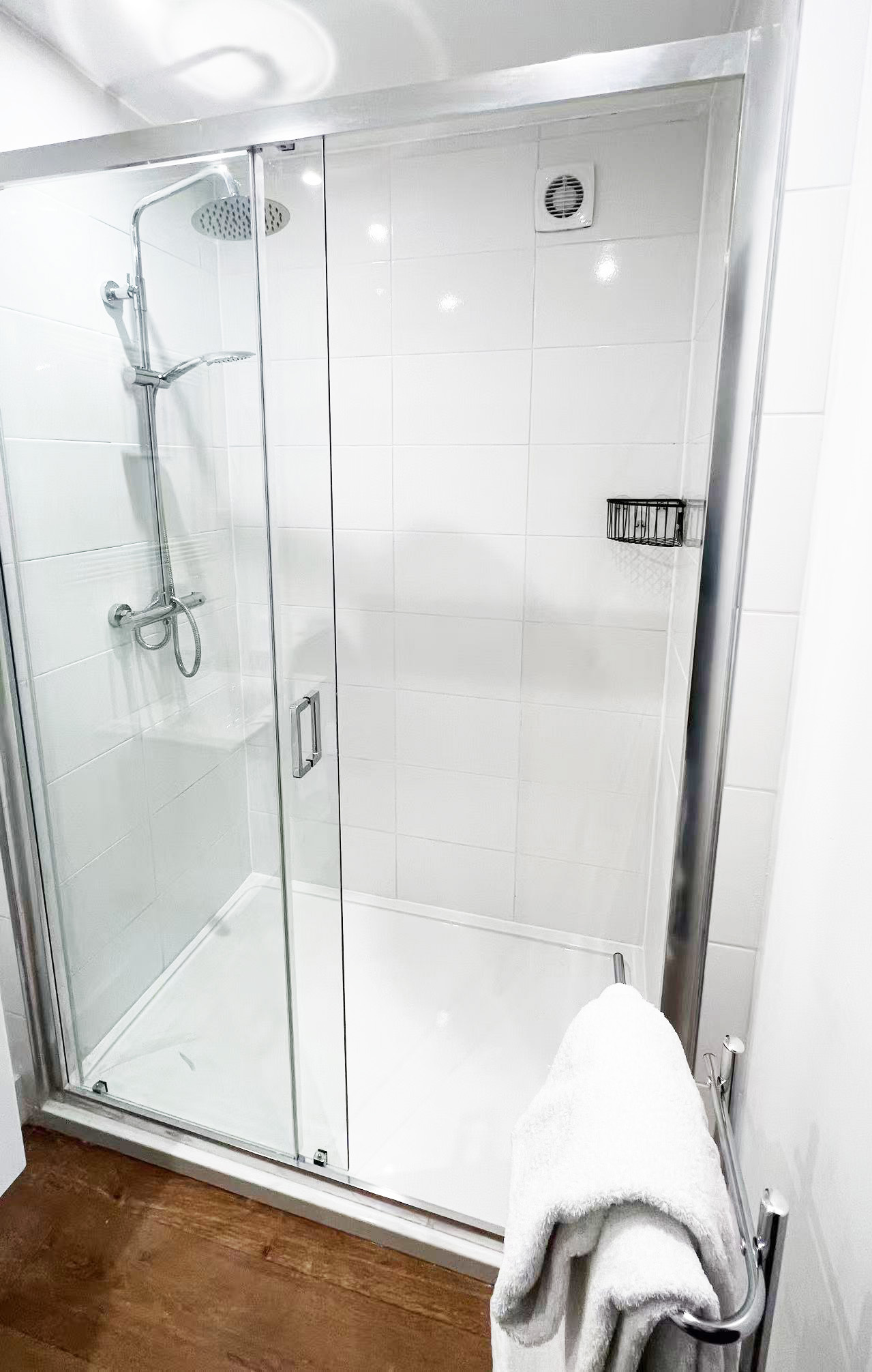 Glass shower enclosure with sliding doors, white tiled walls, and chrome fixtures, including a rain showerhead and handheld shower, seen from the bathroom doorway with a towel on a rack nearby.