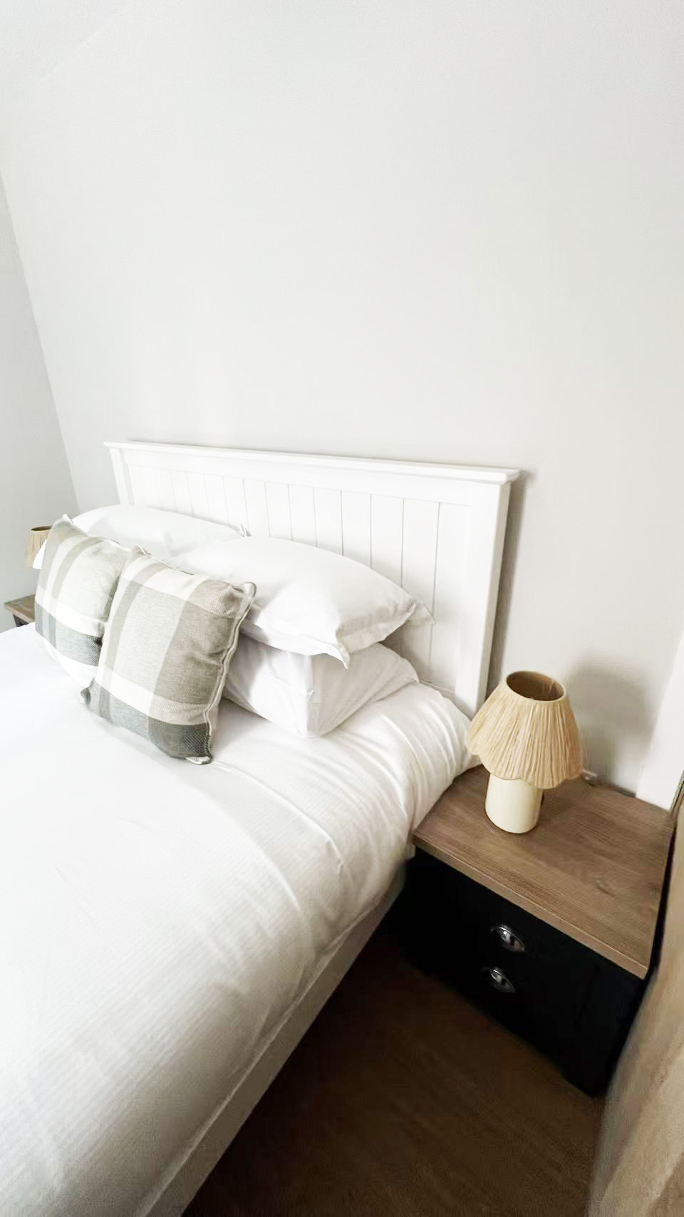 White bed with white linens and gray plaid pillows, a white headboard, and a wooden nightstand with a beige lampshade on the right in a bright, minimal bedroom.