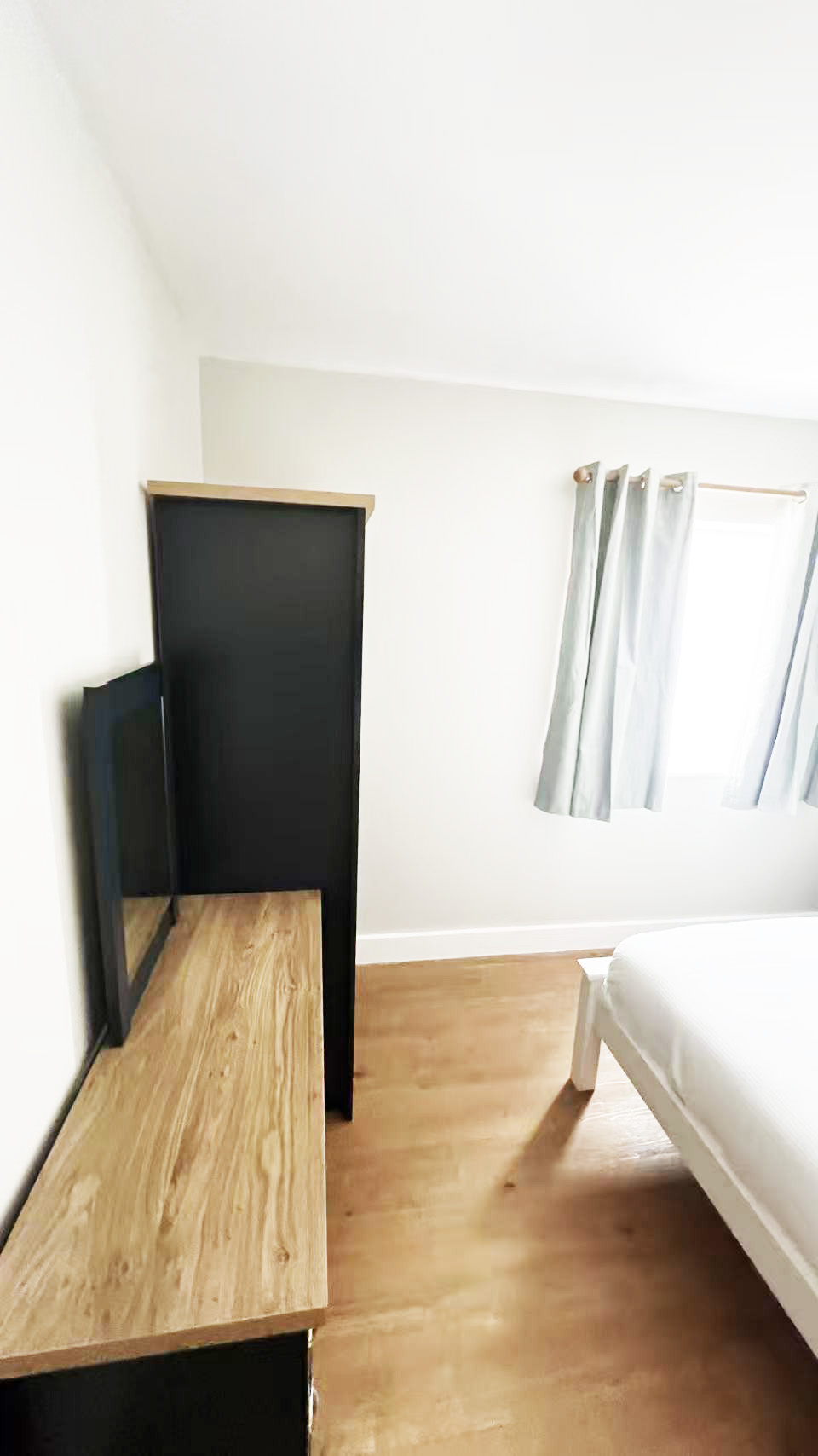 Bedroom scene with a white wall, wooden floor, a bed on the right, and a black dresser with a TV on top. window with light curtains to the right.