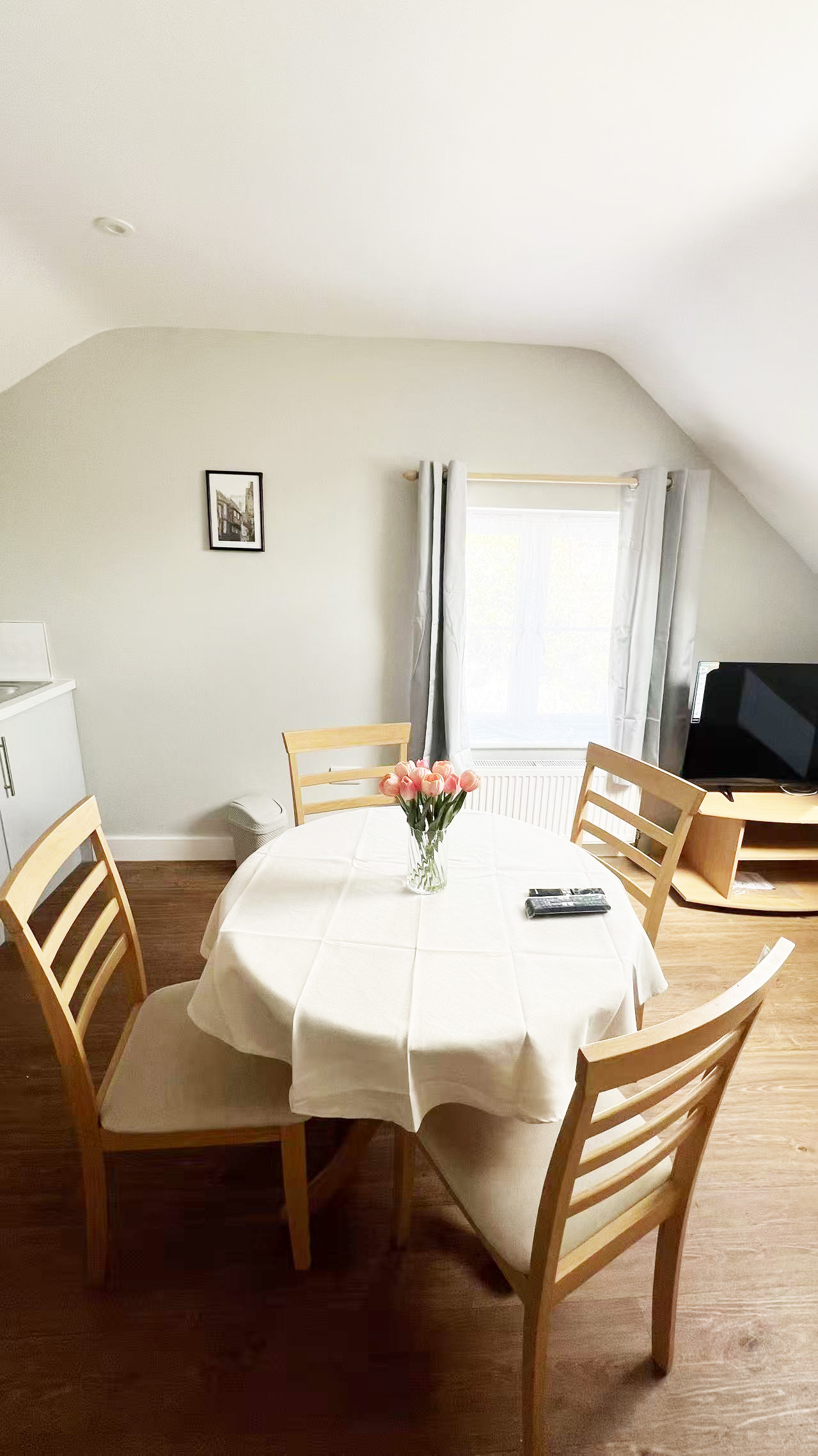 Round dining table with a white tablecloth and a vase of pink tulips, surrounded by wooden chairs in a bright dining nook by a window with light curtains.