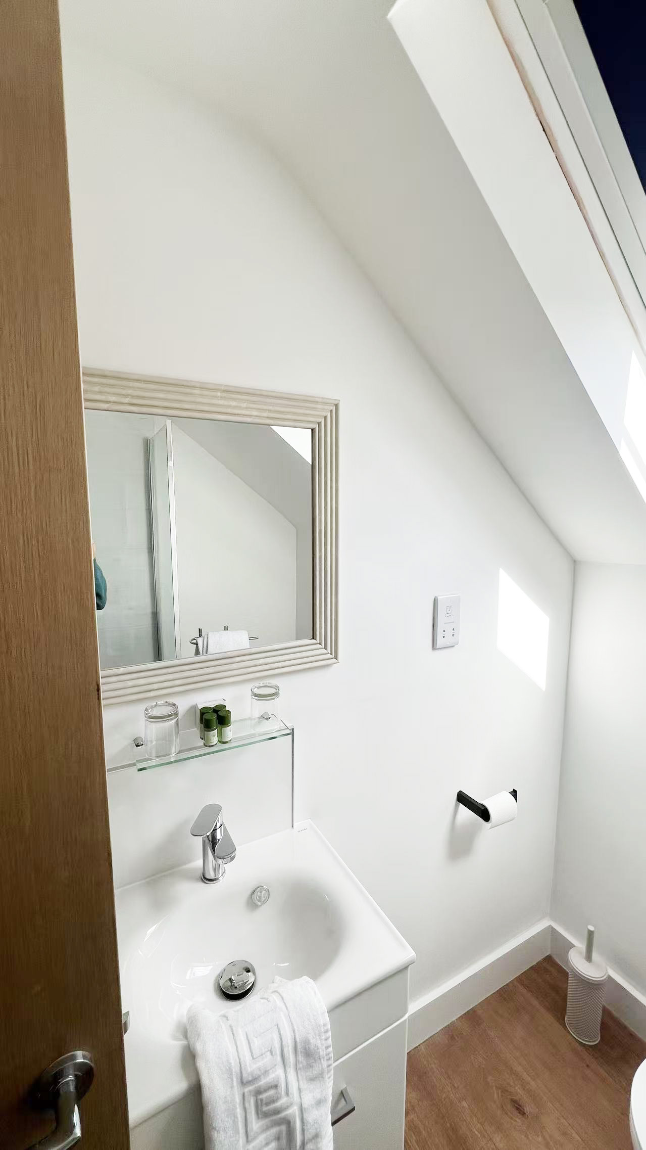 Bright, compact bathroom with white vanity, chrome faucet, towel on the sink, framed mirror, glass shelf with toiletries, and wood-look flooring.