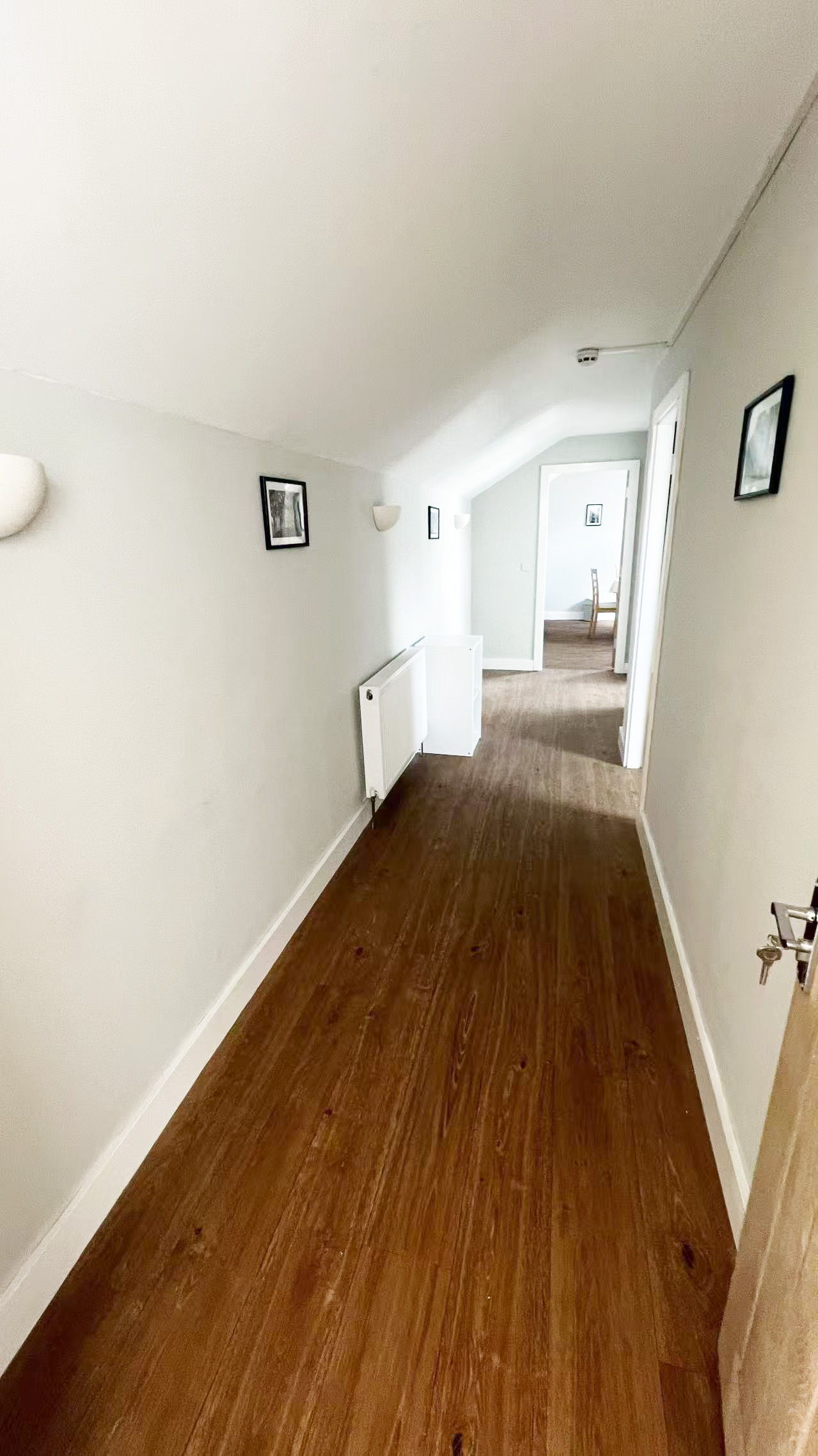 Narrow white hallway with a wooden floor, angled ceiling, and small framed photos on the walls beside a radiator.