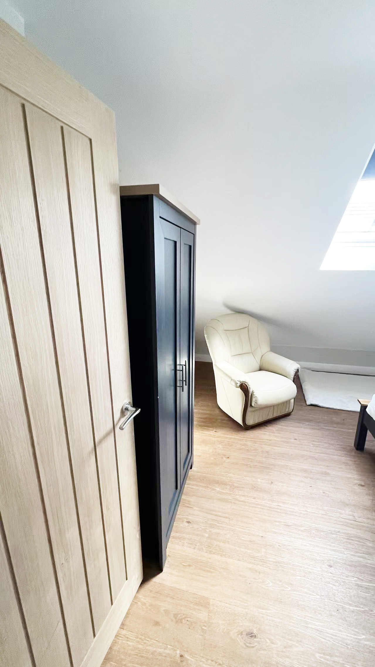 Bright attic corner with a cream leather armchair, a tall dark cabinet, and light wood flooring under a slanted ceiling and skylight.