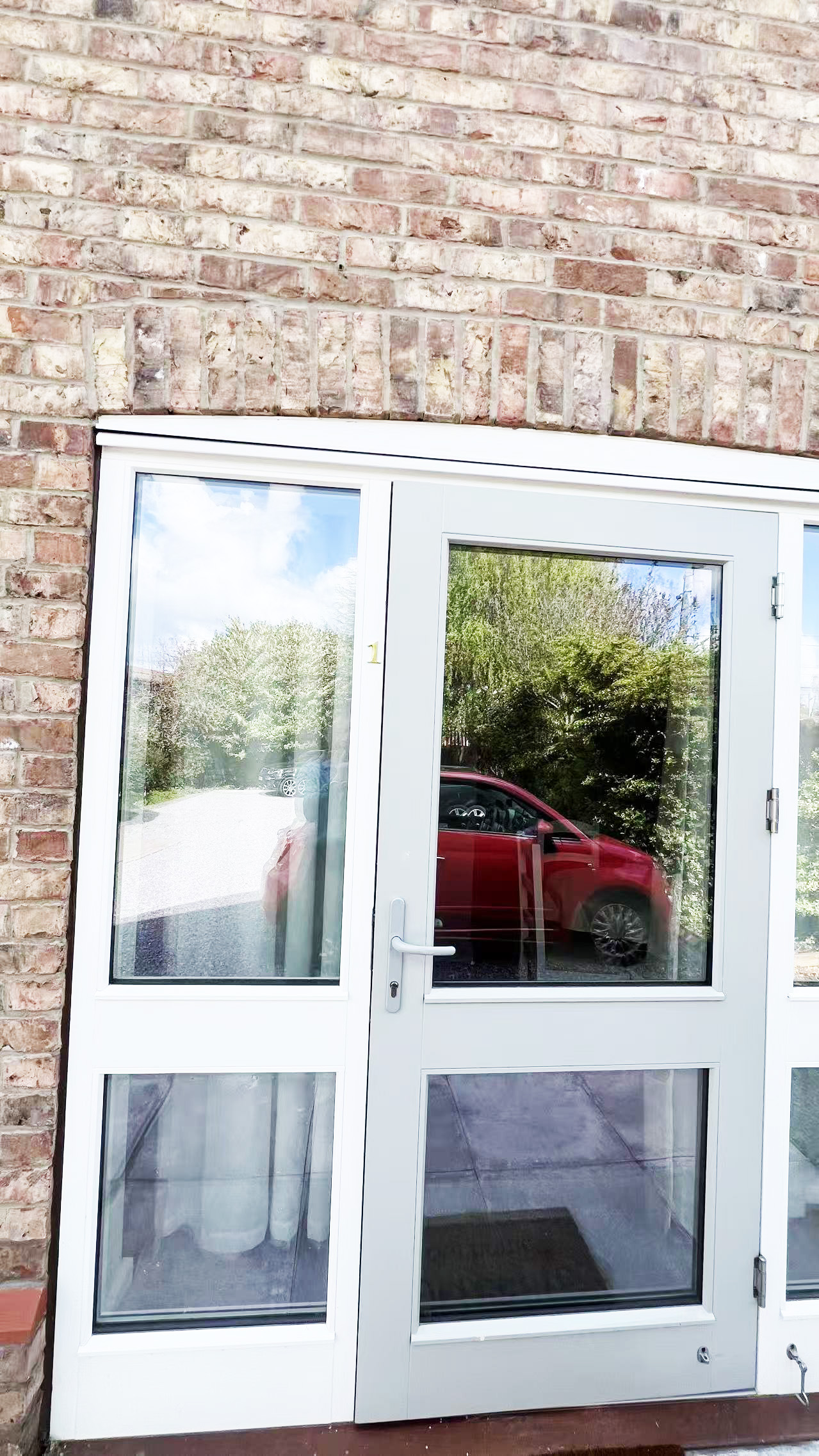 White framed glass door in a brick wall, reflecting trees and a red car outside.