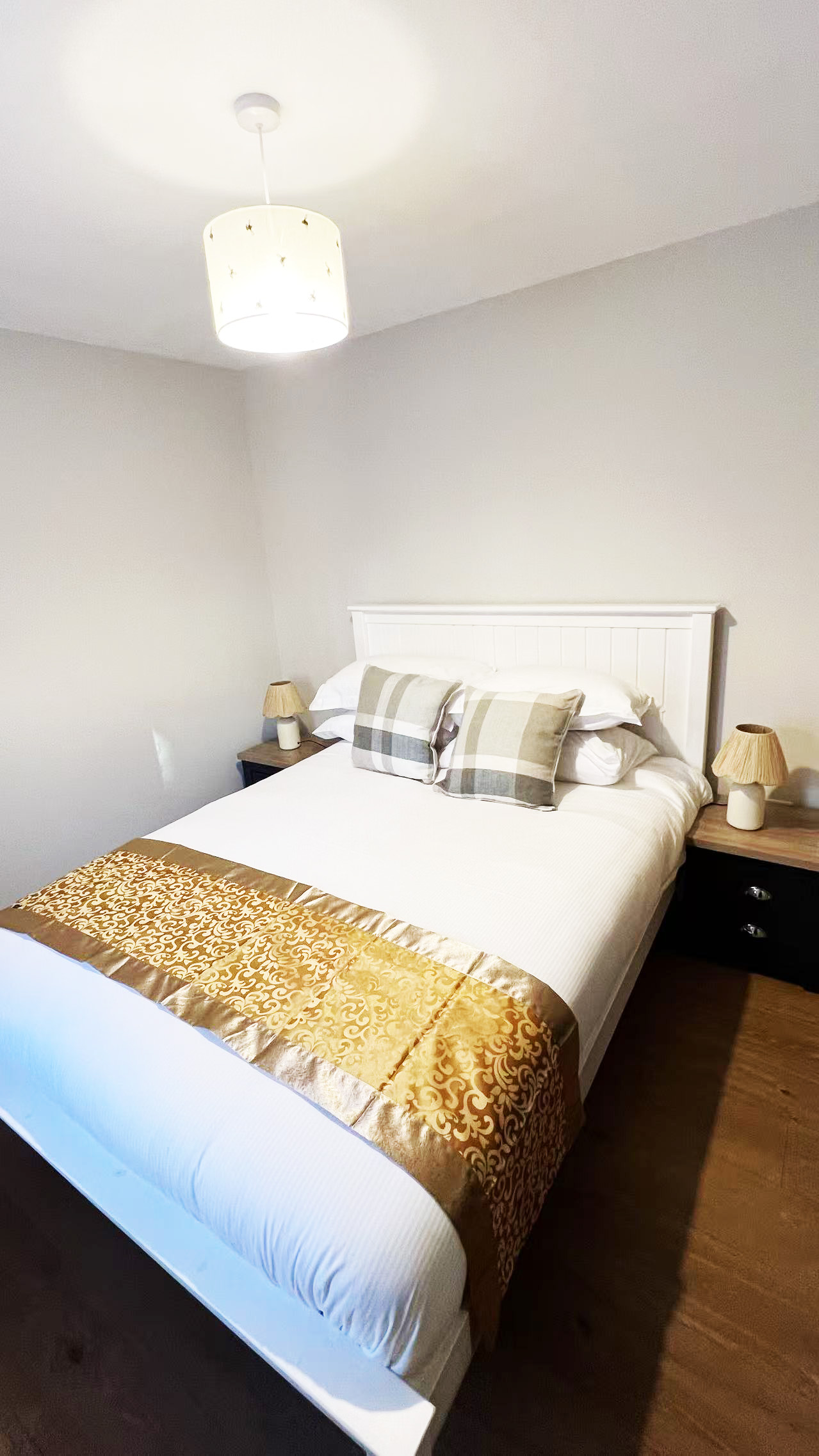 White bedroom with a double bed, plaid pillows, and gold patterned bed runner; two nightstands with lamps flank the bed in a bright room.