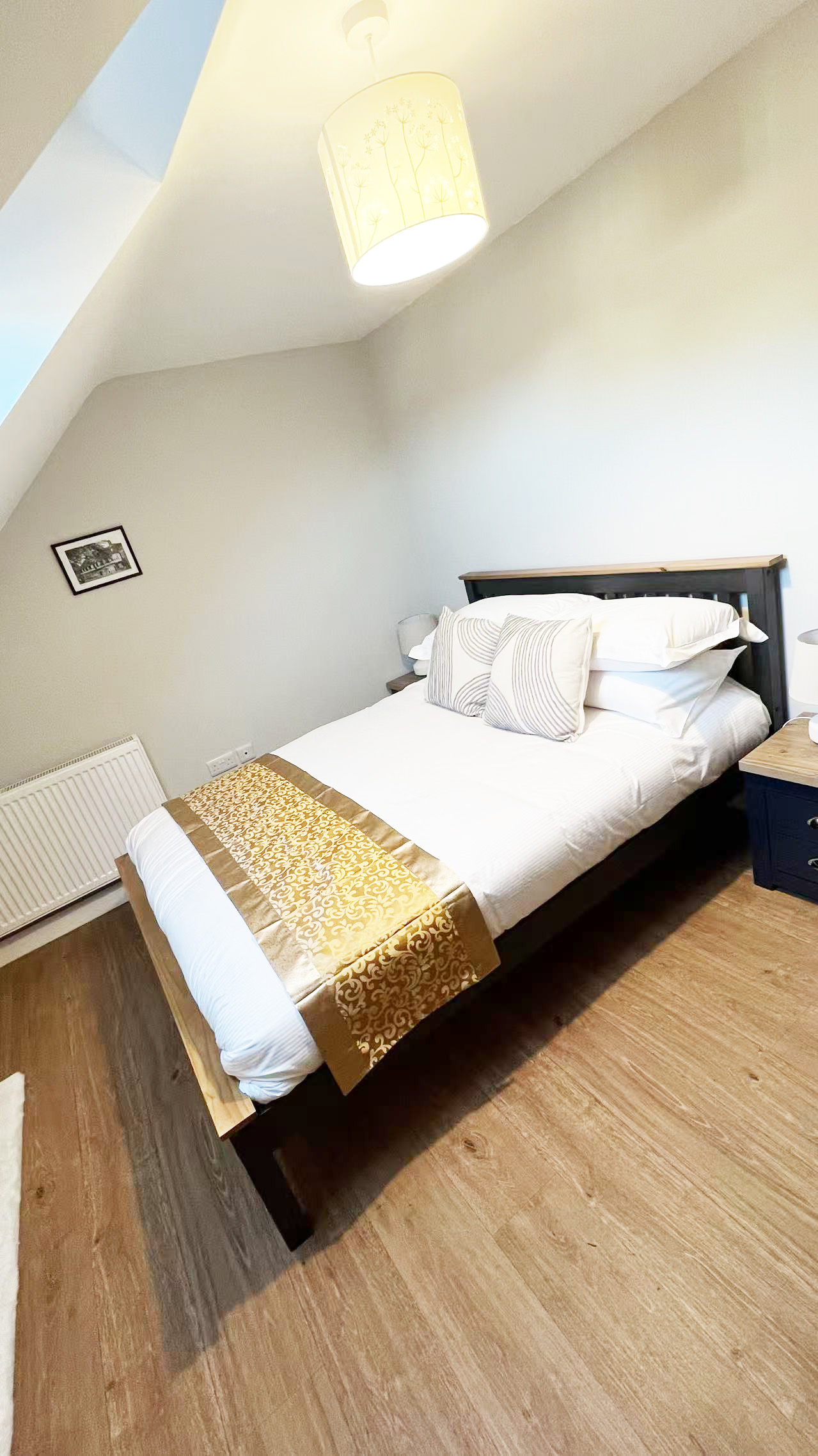 Single bed with white bedding and gold patterned runner, flanked by nightstands in a cozy bedroom under a warm pendant light.