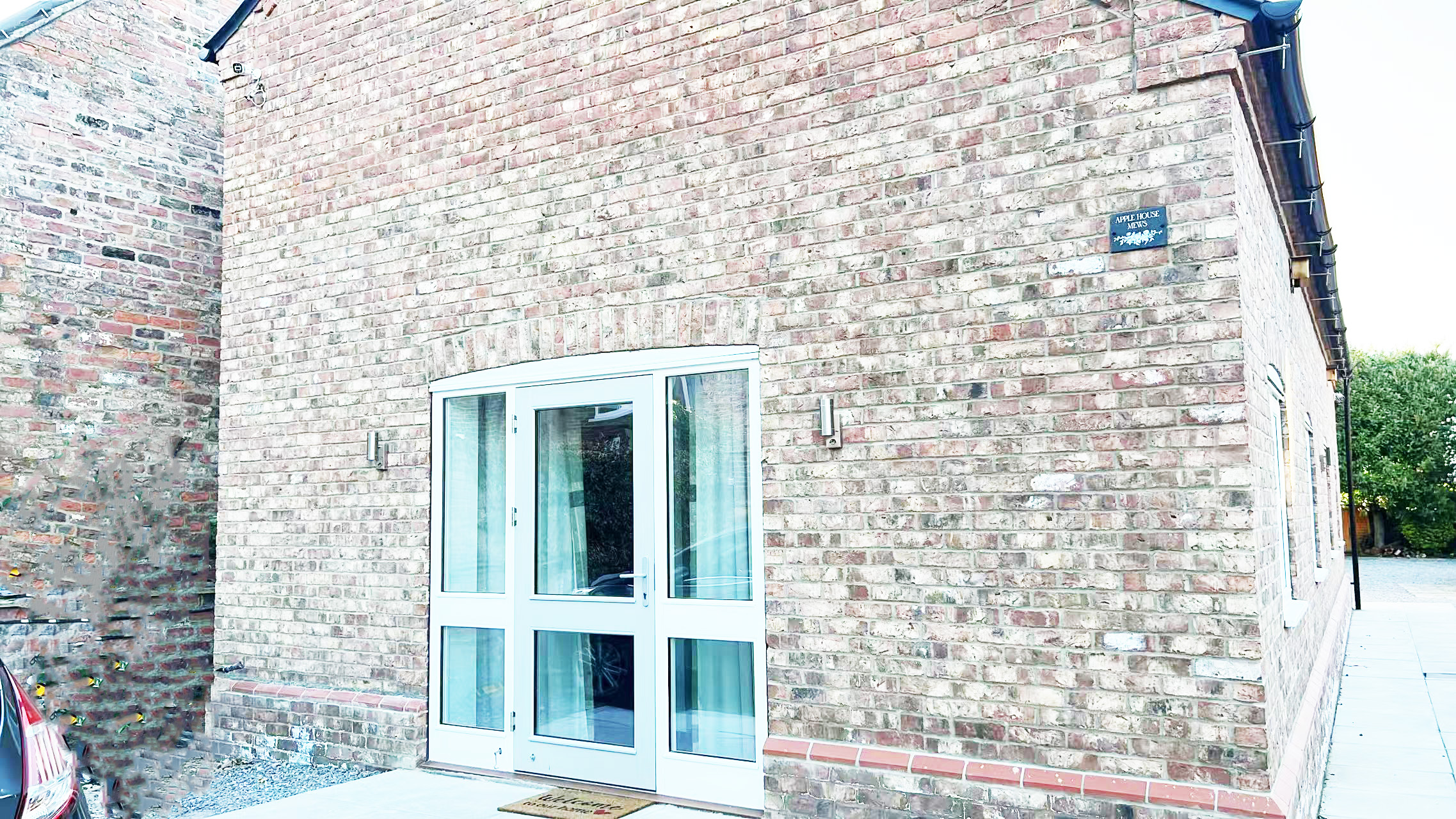 Brick house corner with white double doors and a small welcome mat; blue plaque on the wall reads 'Apple House Mews'.