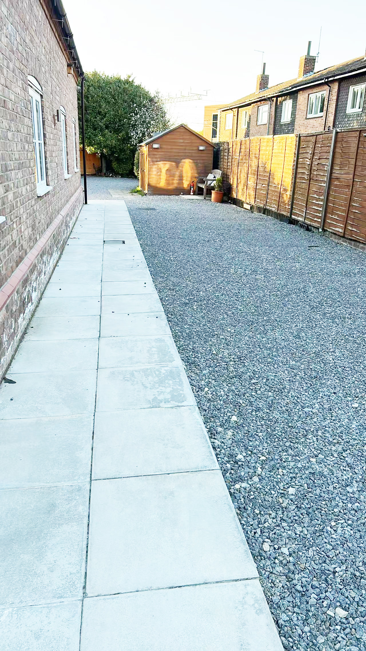 Residential alley with a gravel path, concrete walkway on the left, and a wooden shed at the end amid a wooden fence on the right.