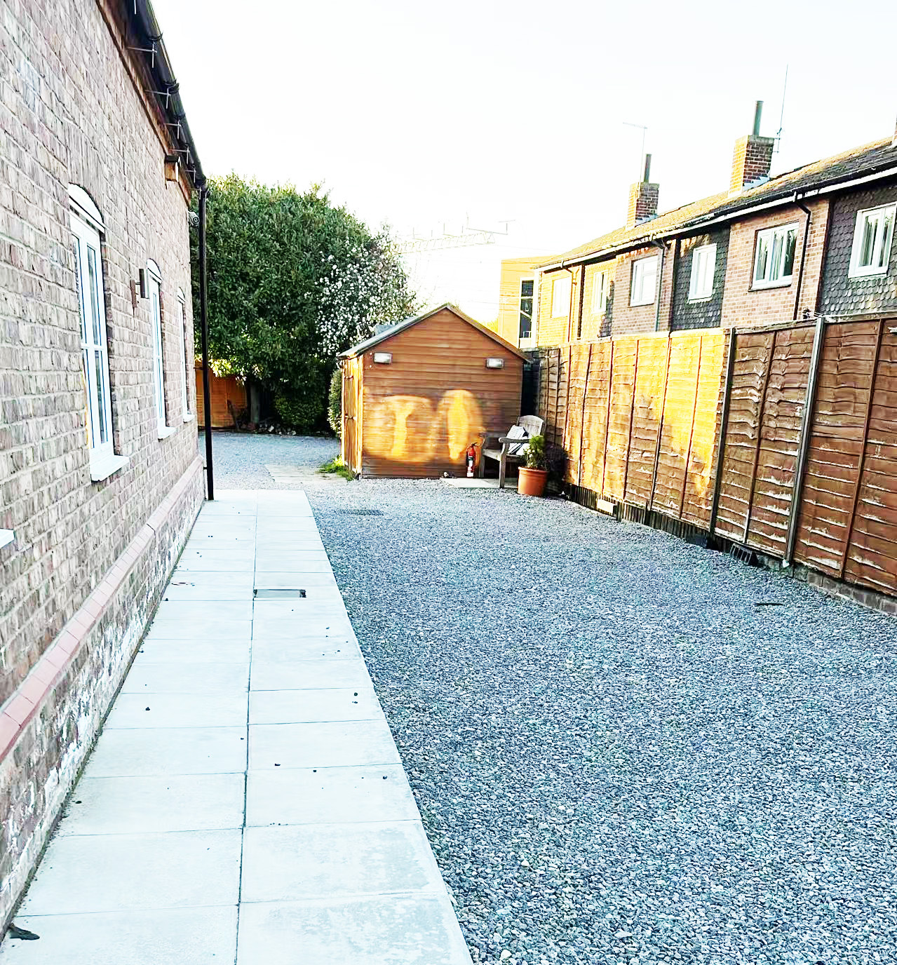 Gravel driveway between brick houses leading to a wooden shed, with a chair and potted plant beside it.
