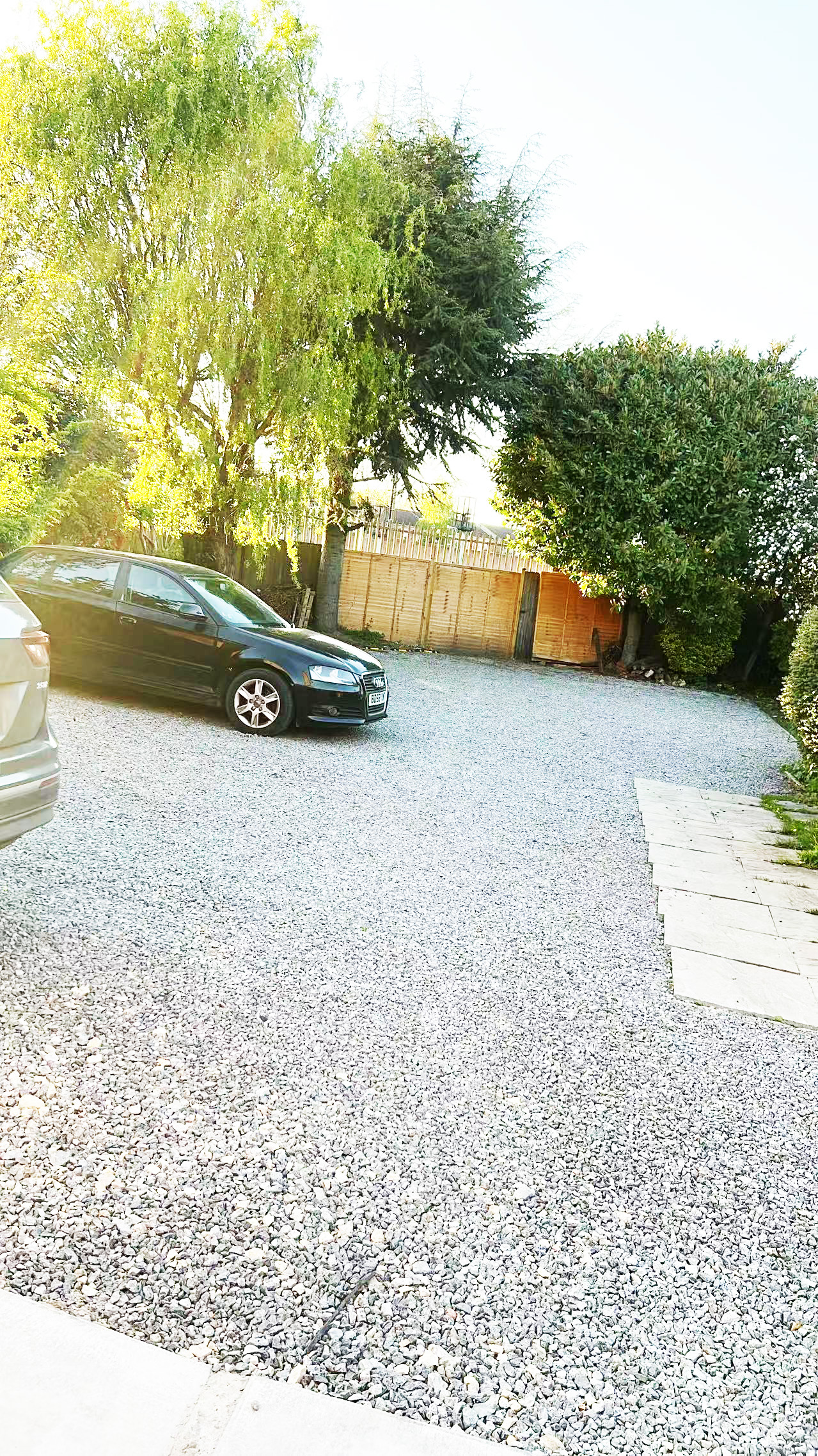 Black sedan parked on a gravel driveway with a wooden fence and tall green trees in the background, bright daylight.
