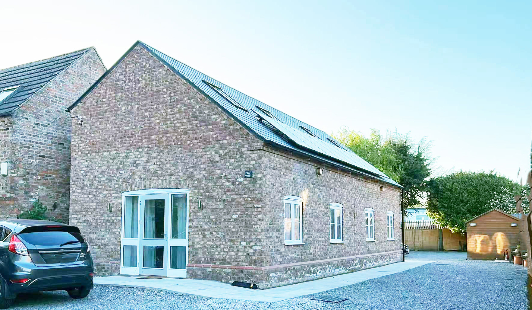 Brick building with a sloped roof covered in solar panels, a blue car parked at the side, and a gravel driveway under a clear sky.