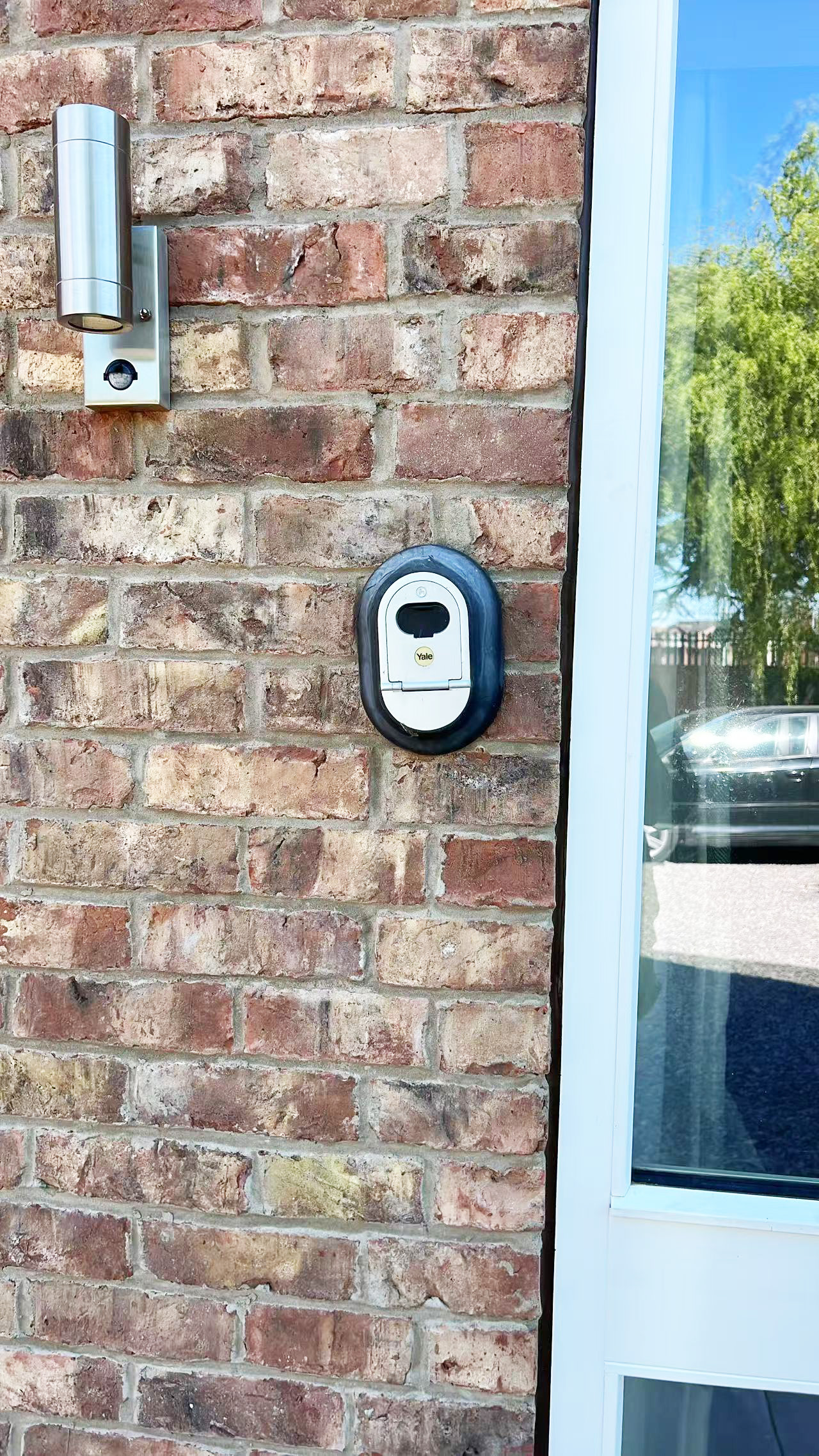 Brick exterior wall with a cylindrical stainless steel wall light and a Yale smart door lock mounted beside a glass door.